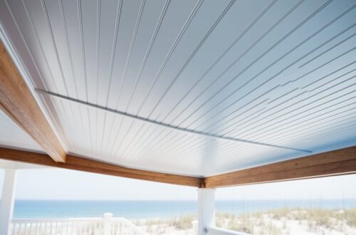 Low-angle view of a coastal porch ceiling finished in white PVC beadboard with a subtle haint-blue tint and hidden fasteners, with white railing, dune grasses, and the ocean softly blurred in the background.