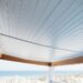 Low-angle view of a coastal porch ceiling finished in white PVC beadboard with a subtle haint-blue tint and hidden fasteners, with white railing, dune grasses, and the ocean softly blurred in the background.