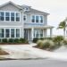 Street-level view of a coastal home exterior with power-washing focused on the driveway and refreshed native landscaping, with a blurred ocean view in the background.