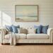 Sunlit traditional coastal living room with a cream tufted sofa, sisal rug, rattan side tables, and framed art above whitewashed shiplap.