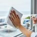 Hands wiping a clear window with a microfiber cloth and spray bottle in a bright coastal home with an ocean view outside.