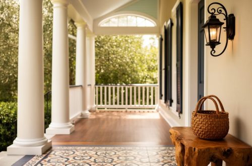 Sunlit Southern porch and interior with white columns, wrought-iron decor, indigo textiles, and a sweetgrass basket on a cypress table, suggesting diverse regional craftsmanship.